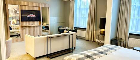 A hotel room with a large bed, a TV mounted on the wall, a seating area with a sofa and chairs, and a small table.
