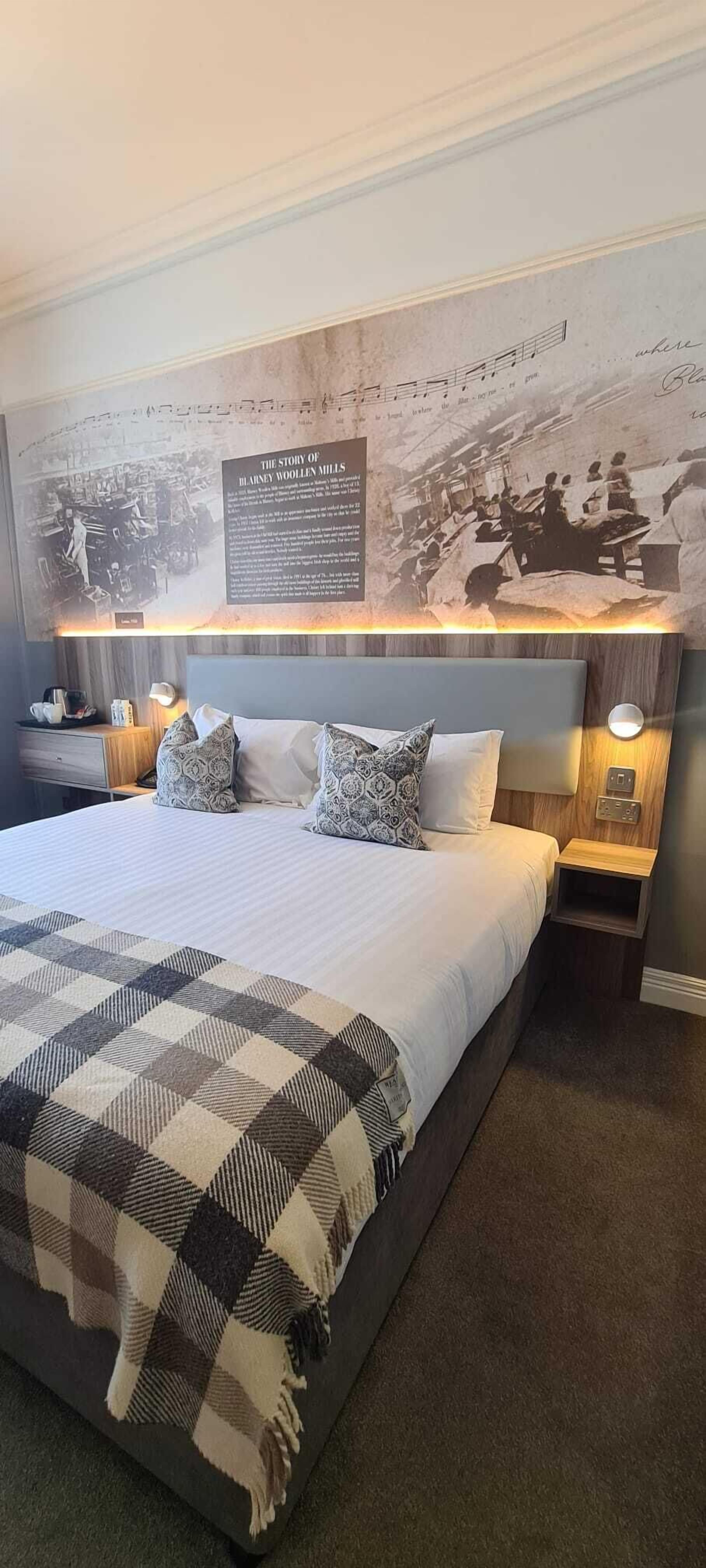 Una habitación de hotel con una cama grande, un cabecero con una fotografía histórica, lámparas de noche y una manta de cuadros.