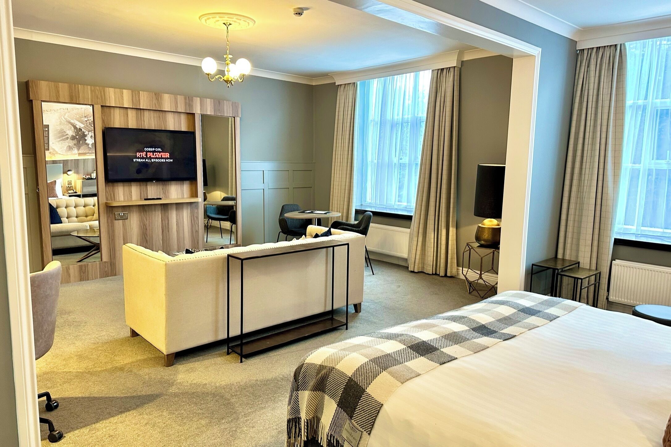 A hotel room with a large bed, a sofa, a desk, a television, and a window with curtains.