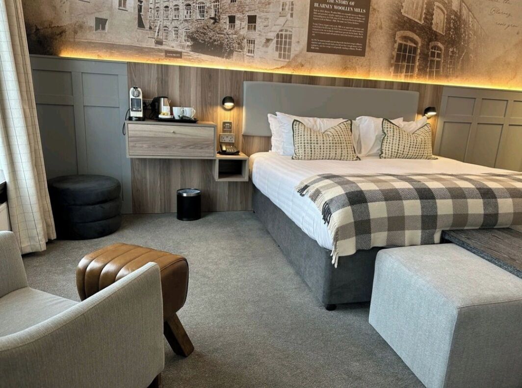 Una habitación de hotel moderna con una cama grande, una mesita de noche de madera, una silla, un pequeño otomaní y un estante montado en la pared.