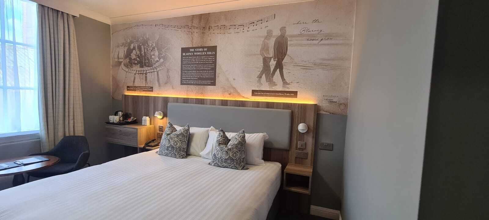 A hotel room with a large bed, a desk, and a chair. There are two pillows on the bed and a lamp on the desk.