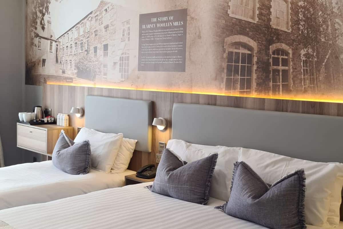 Habitación de hotel con dos camas, cada una con cabecero y almohadas, una mesita de noche con teléfono y una lámpara de pared.
