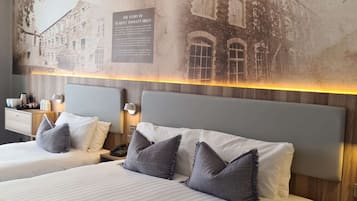 Habitación de hotel con dos camas, cada una con cabecero y almohadas, una mesita de noche con teléfono y una lámpara de pared.