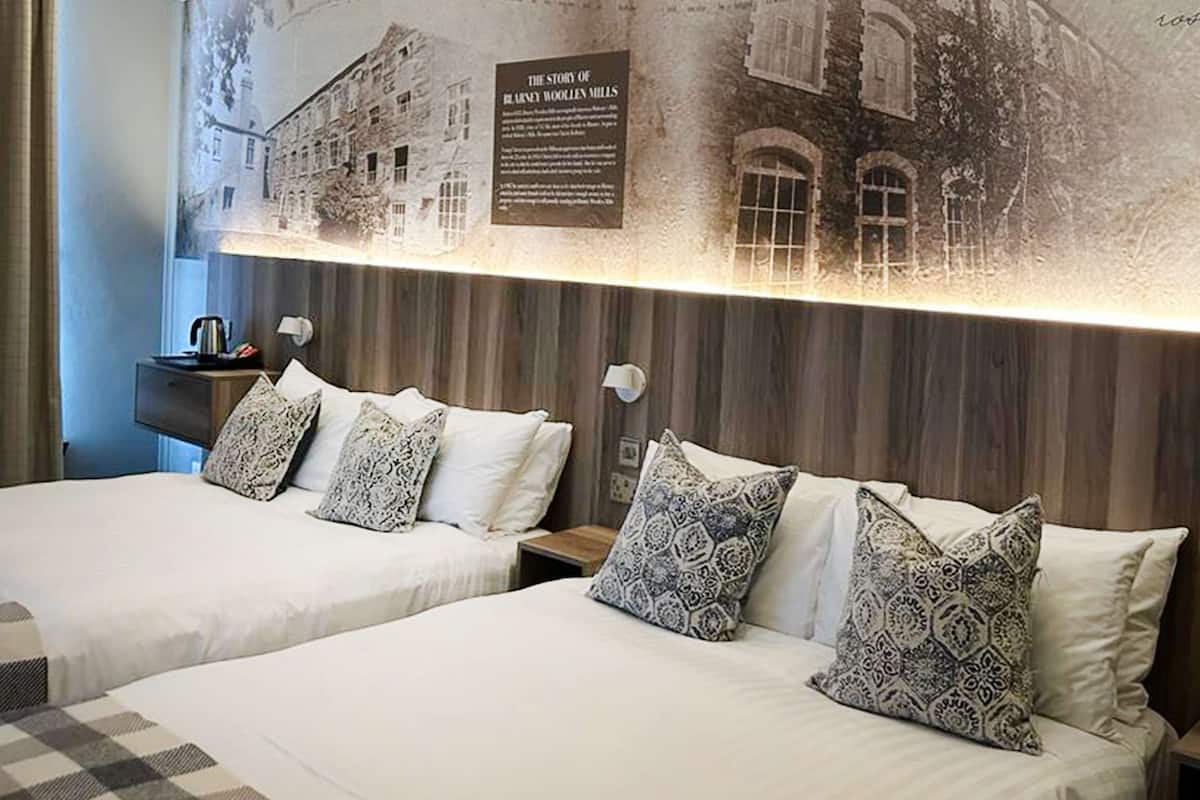 Una habitación de hotel con dos camas, almohadas estampadas, una lámpara fijada a la pared y una placa conmemorativa histórica.