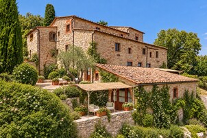 Un edificio de piedra con tejado de baldosas, rodeado de abundante vegetación y un patio con mobiliario exterior.