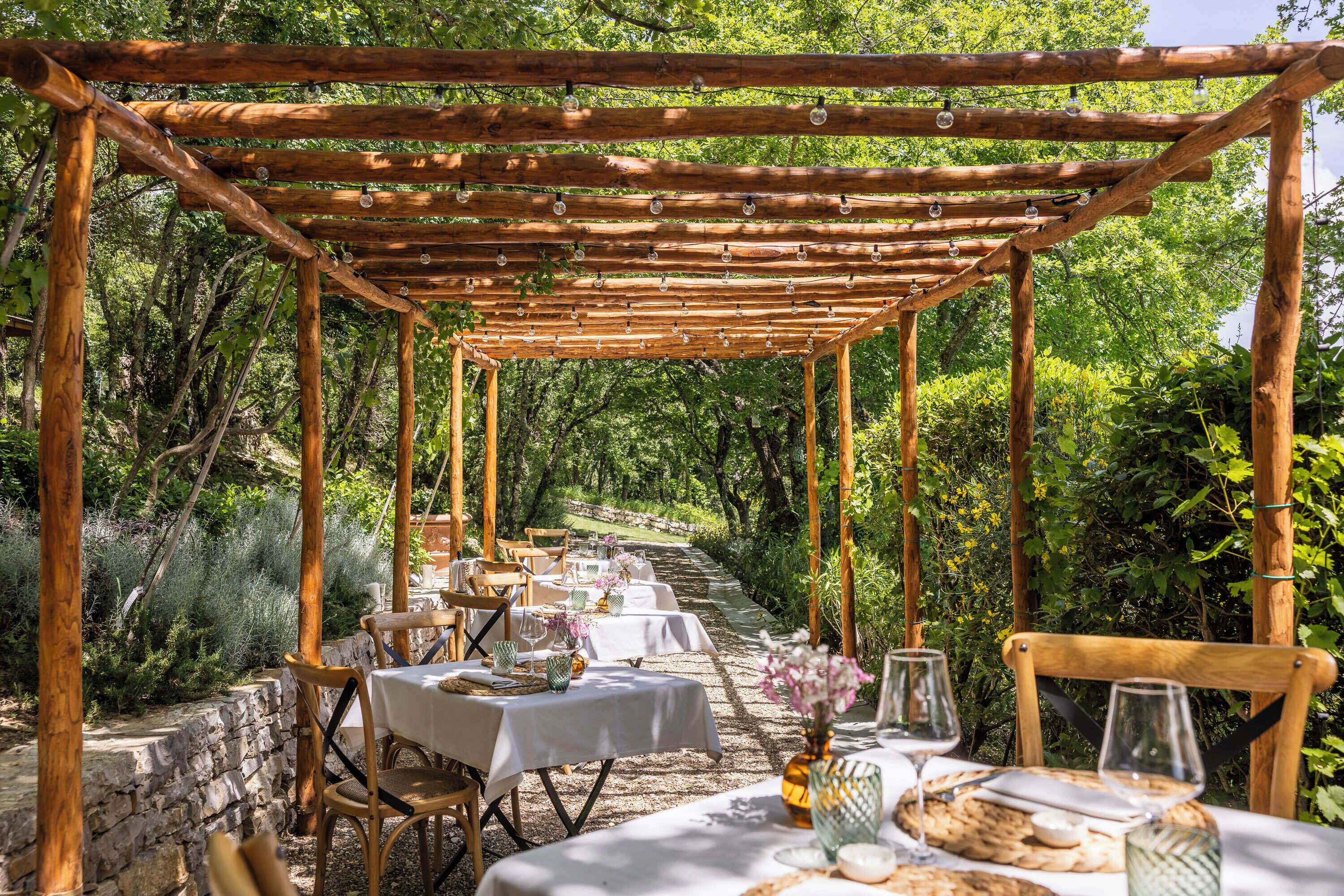 An outdoor dining area with tables set under a wooden pergola, surrounded by greenery.