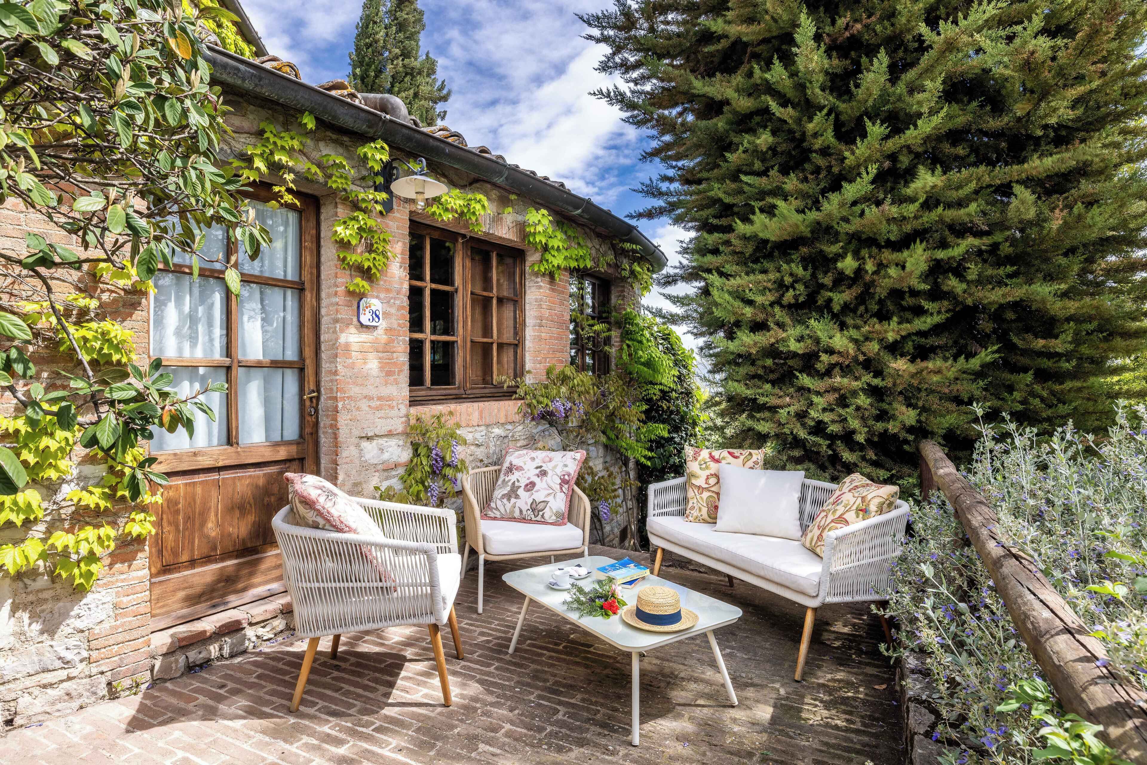 A cozy outdoor seating area with wicker chairs and a white sofa, a brick building with ivy, and a large evergreen tree.