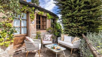 A cozy outdoor seating area with wicker chairs and a white sofa, a brick building with ivy, and a large evergreen tree.
