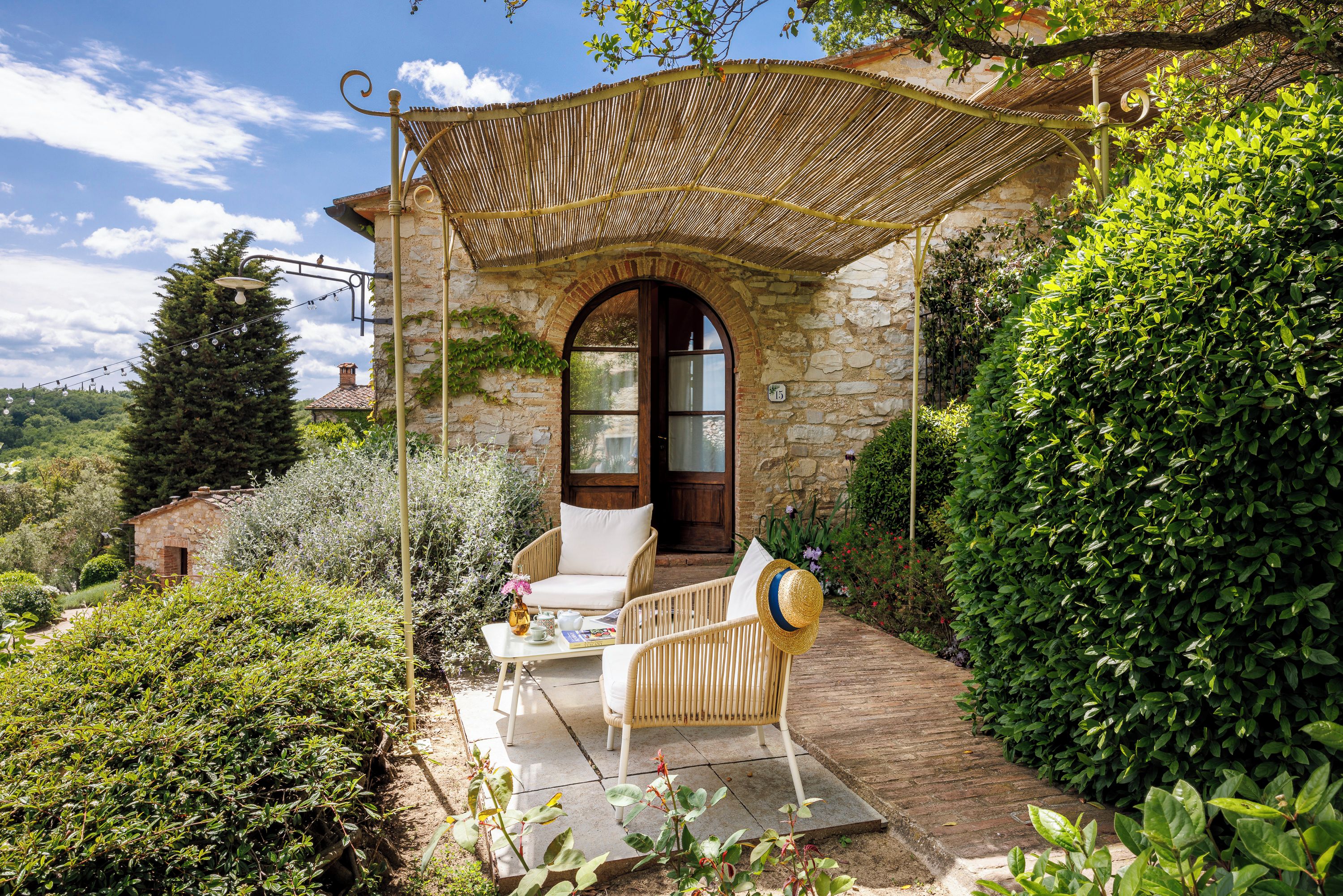 A stone building with a covered patio, a wooden chair, a table with a vase and a book, and a large bush.