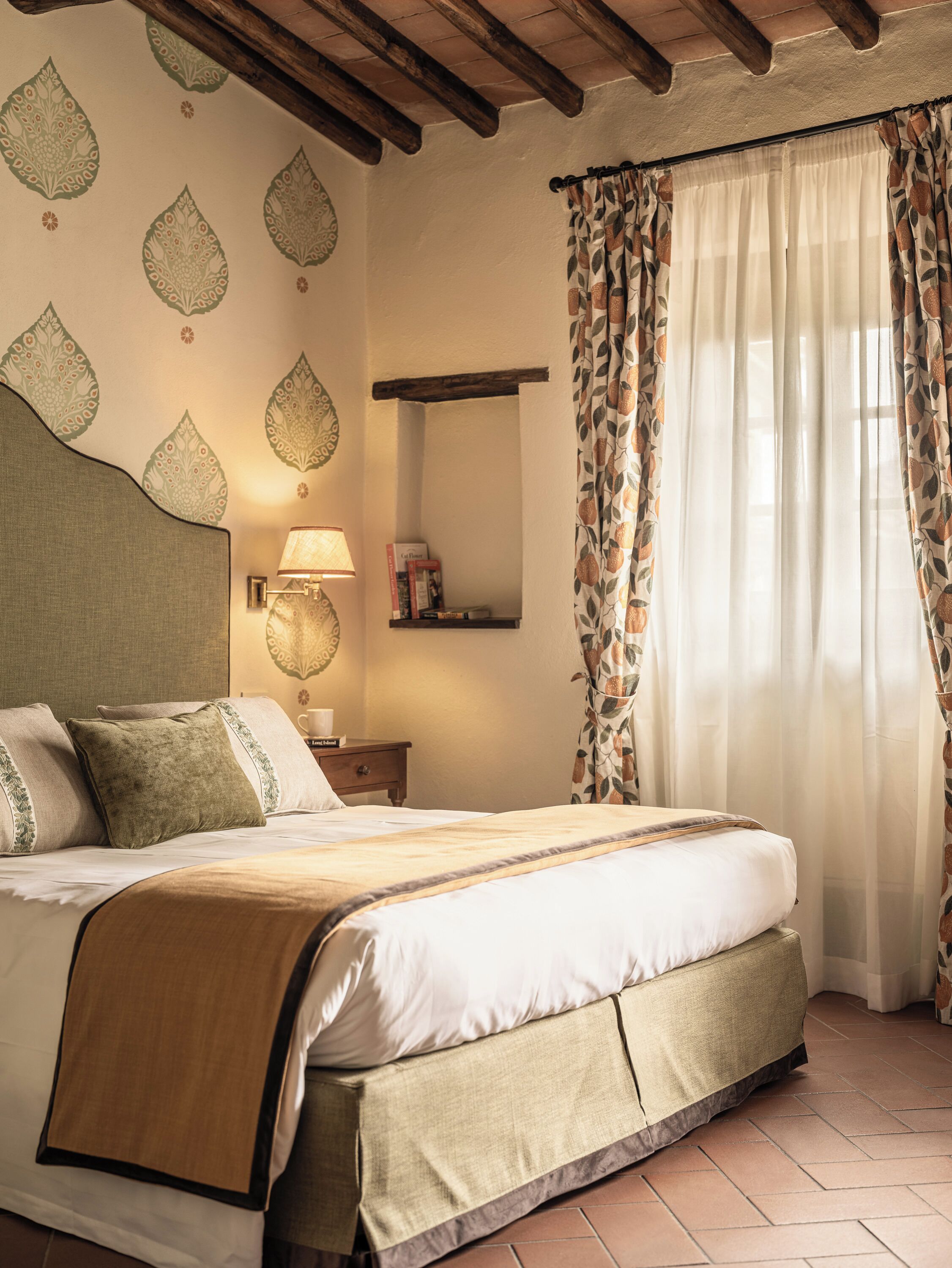 Une chambre à coucher avec un lit, une table de chevet, une lampe, un miroir et une fenêtre avec des rideaux.