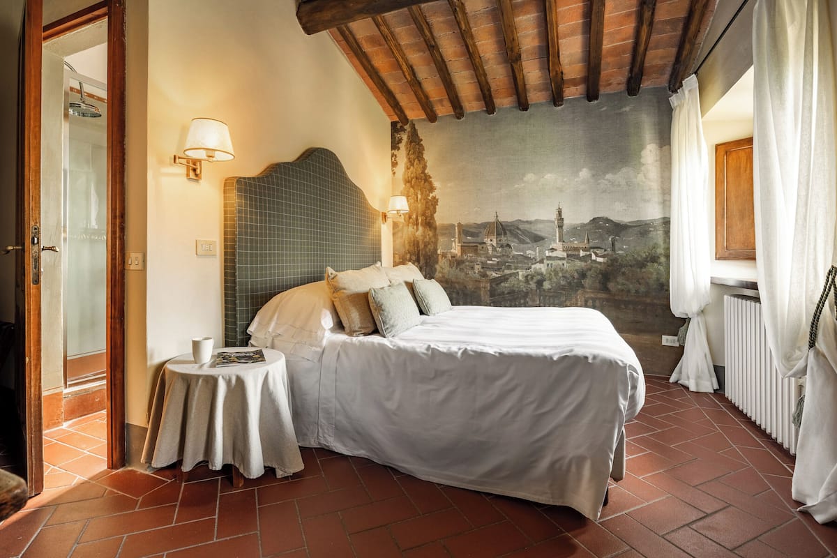 A bedroom with a bed, a small table, a lamp, a mural, and a window with curtains.