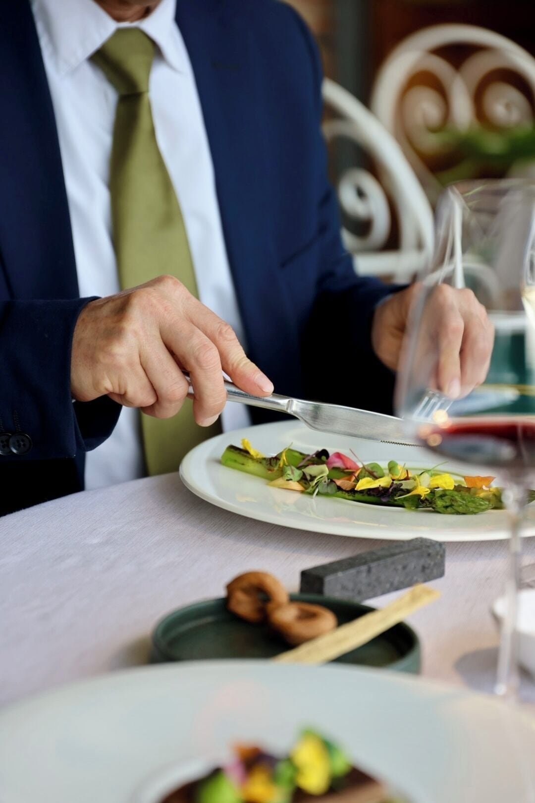 Una persona vestida con traje cena en una mesa con un plato de comida y una copa de vino tinto.