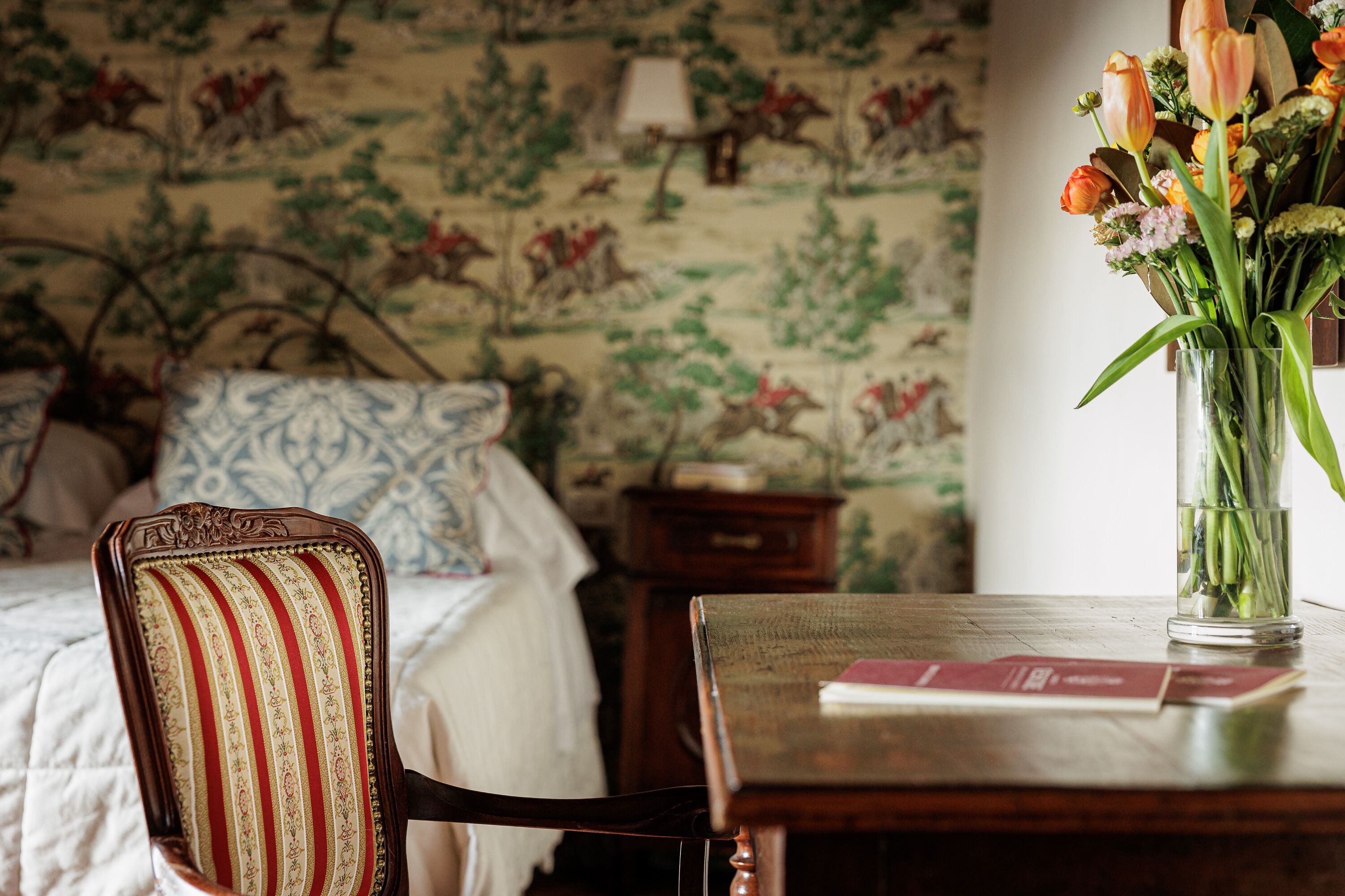 A room with a bed, a chair, a table with a vase of flowers, and a wall with a floral pattern.