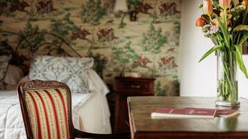 A room with a bed, a chair, a table with a vase of flowers, and a wall with a floral pattern.