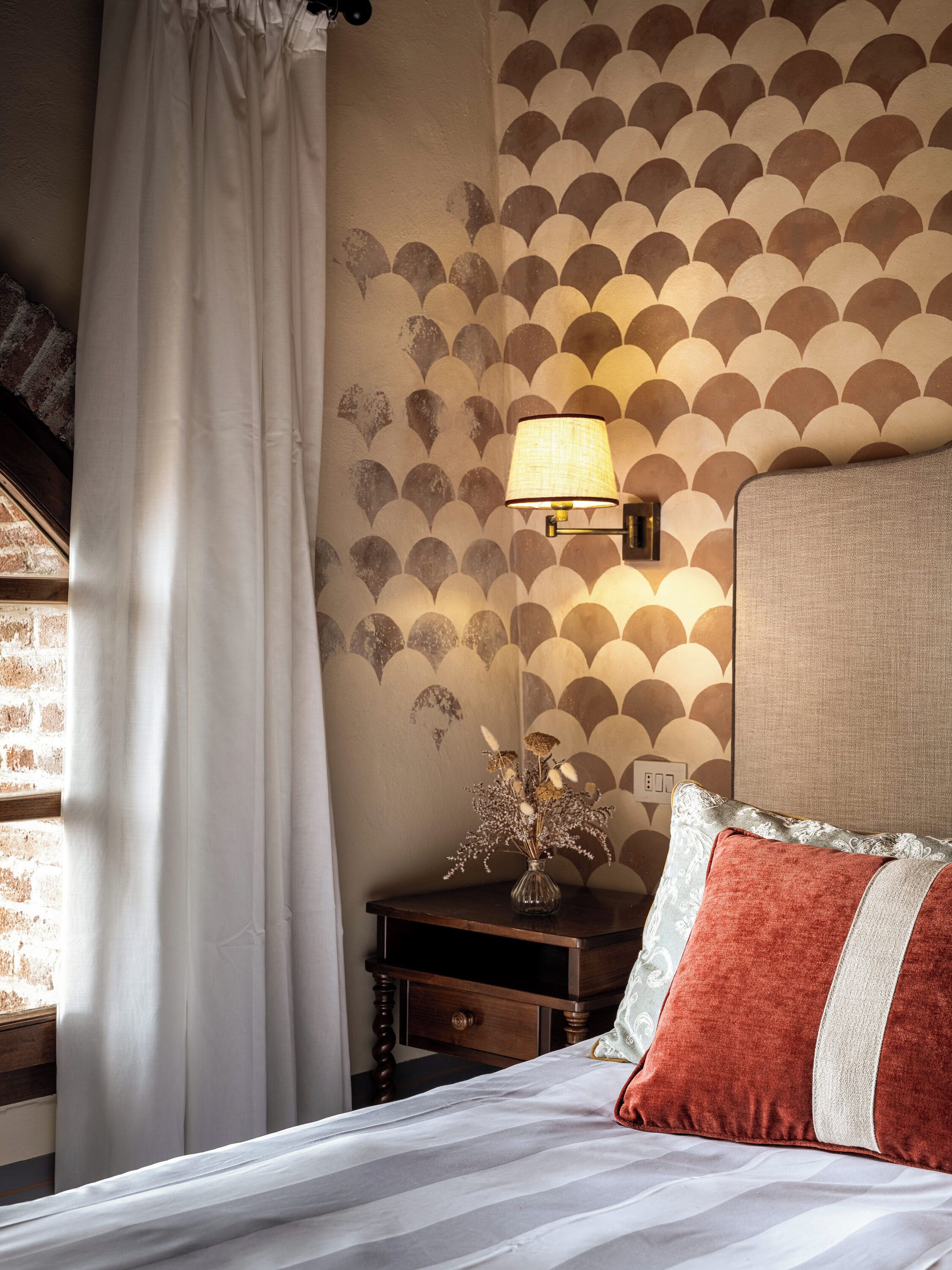 Quarto com cama, mesa de cabeceira, abajur, um vaso com flores secas e parede estampada.