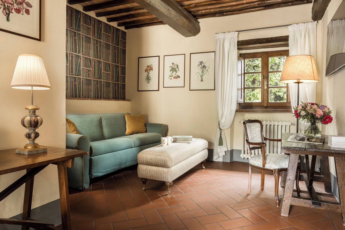 A cozy room with a wooden table, a blue sofa, a cream-colored ottoman, and a chair. There are framed botanical prints on the wall, a radiator, and a window with curtains.