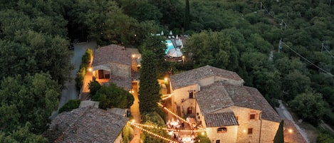 An aerial view of a rustic village with stone buildings, a central courtyard with string lights, and a swimming pool.