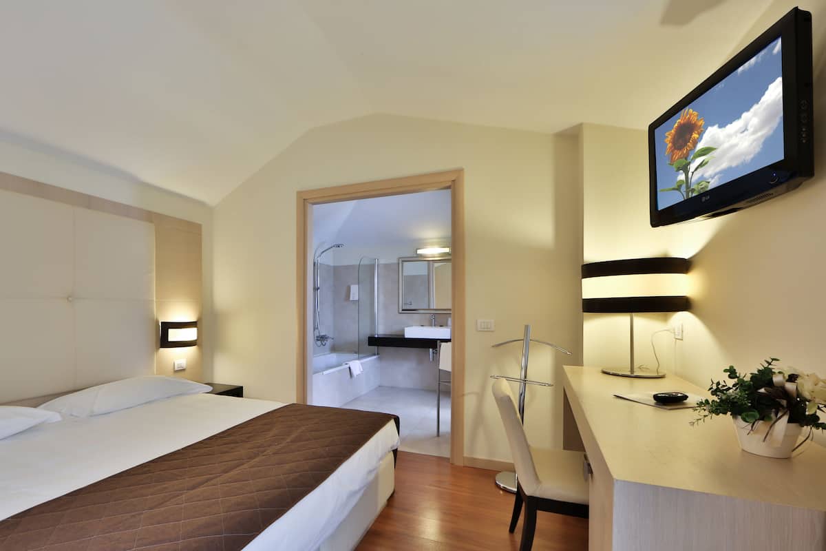 A modern hotel room with a bed, a desk with a chair, a bathroom with a shower and sink, and a large window with a view of the sky and clouds.
