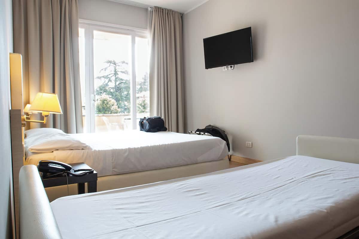 A hotel room with two beds, a TV on the wall, a window with curtains, a chair, and a desk with a lamp.