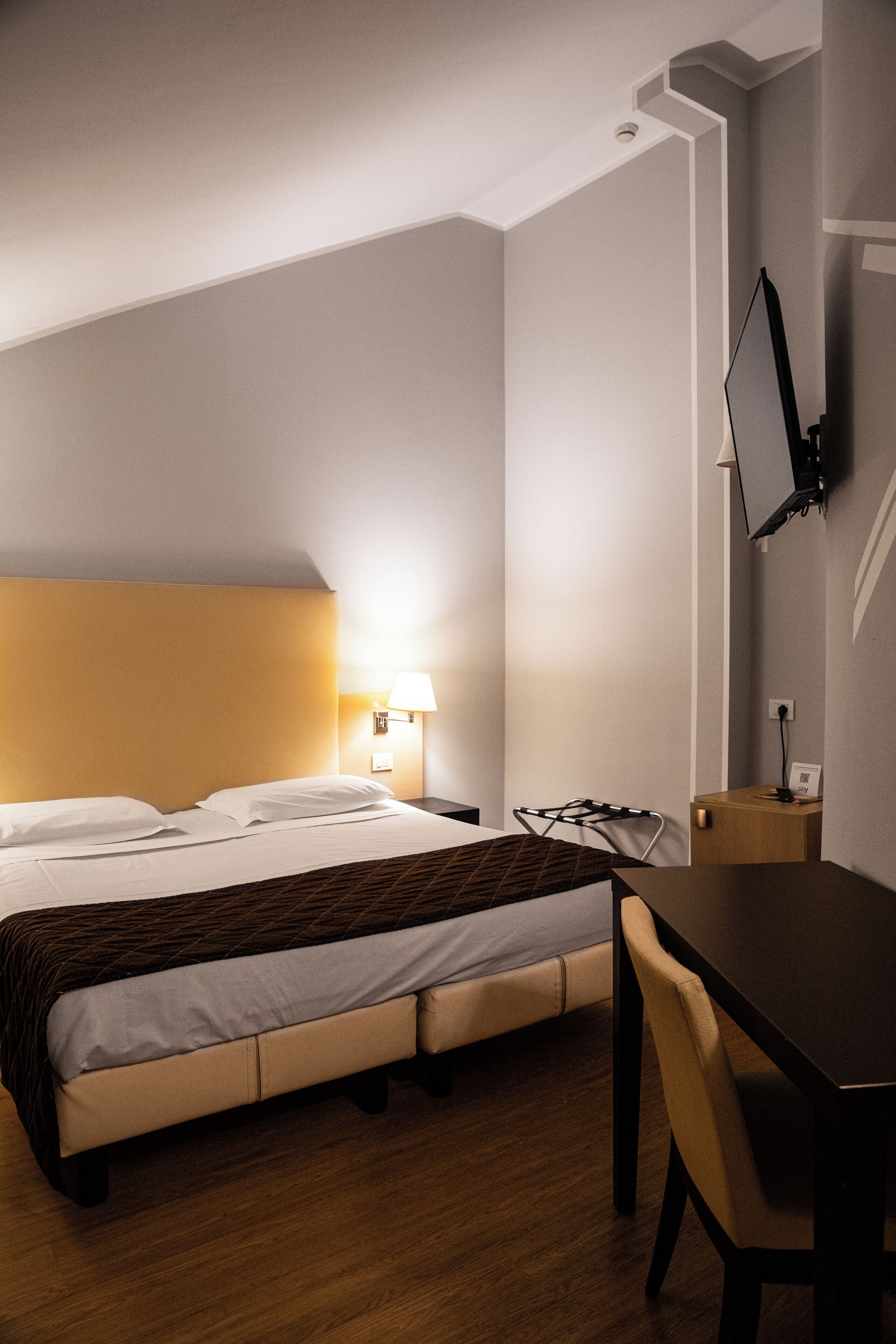 A hotel room with a bed, a desk, a chair, a TV mounted on the wall, and a lamp.