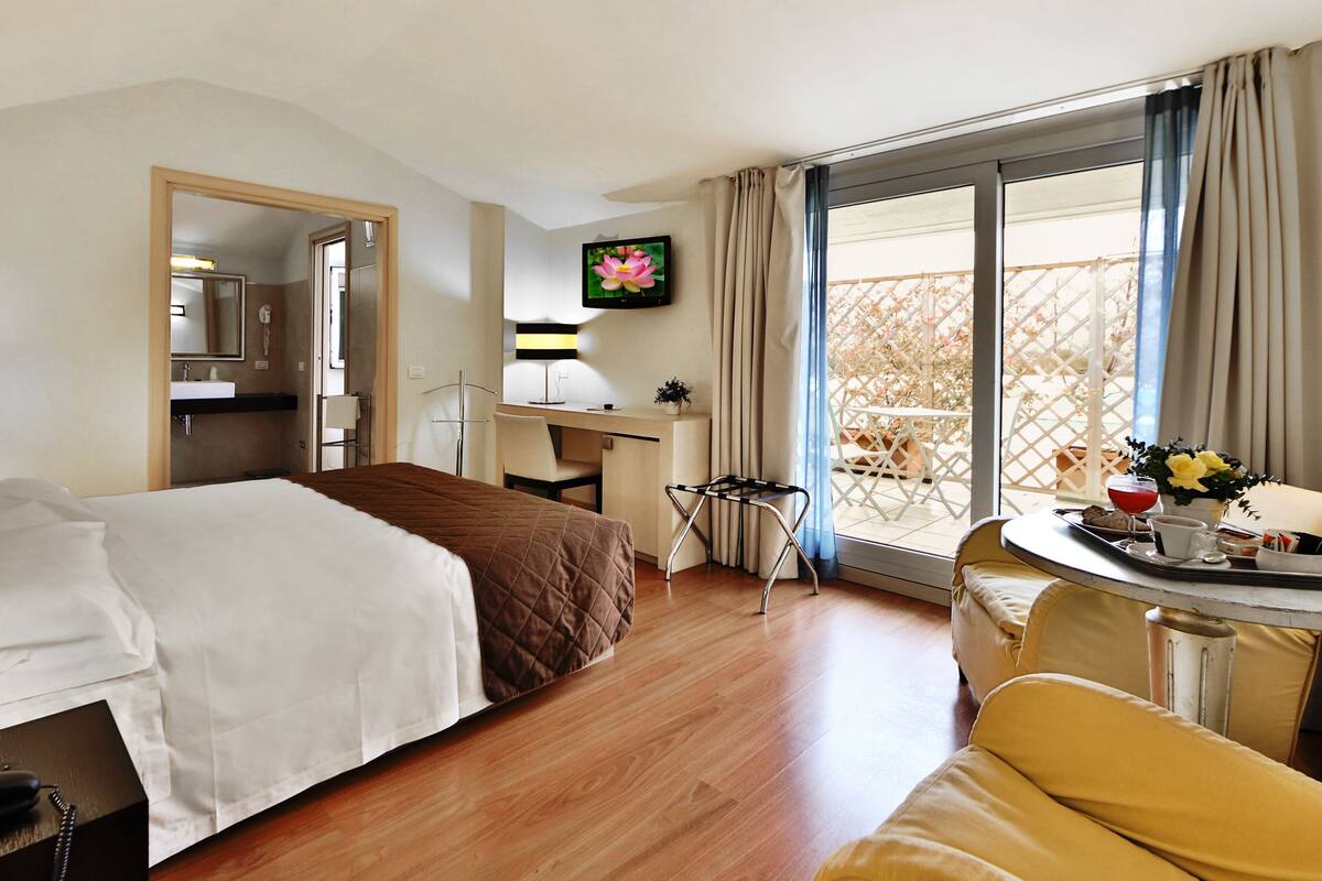 A hotel room with a bed, a desk, a chair, a small table with breakfast items, and a balcony with a view.