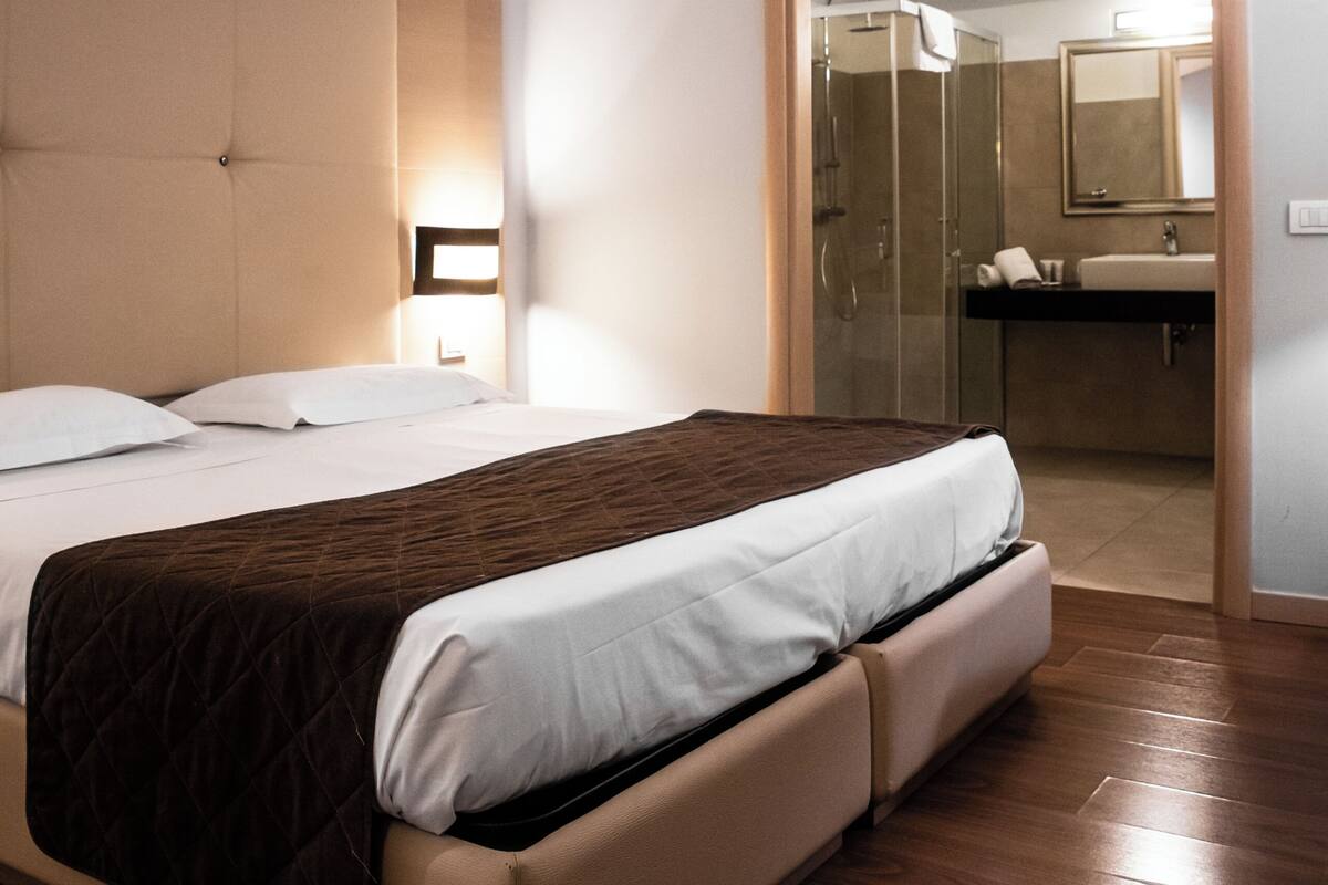 A bedroom with a bed, a bedside table, a mirror, and a bathroom visible through an open door.