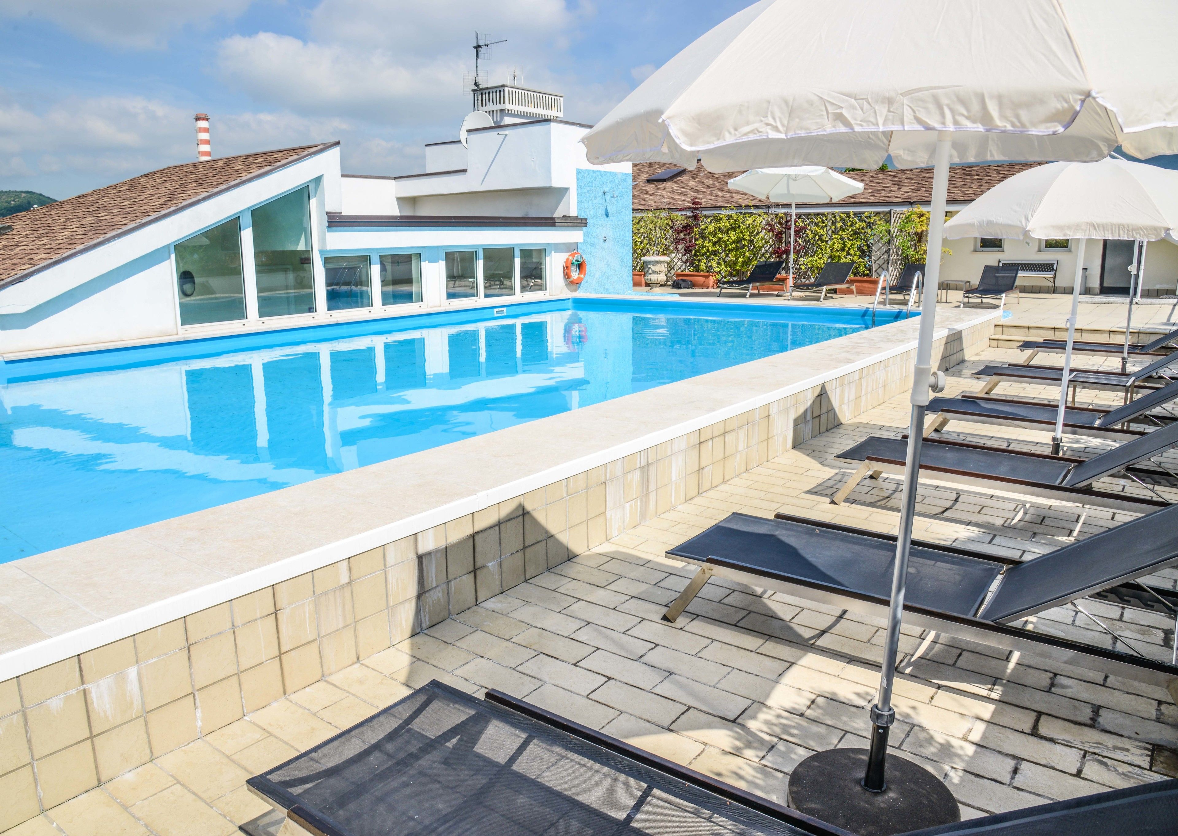 Ein Schwimmbecken mit Liegestühlen und Sonnenschirmen, einem Rettungsring und einem Gebäude mit Satteldach und Schornstein.