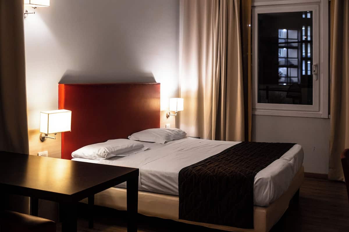 A hotel room with a bed, bedside lamps, a table, and a window with curtains.