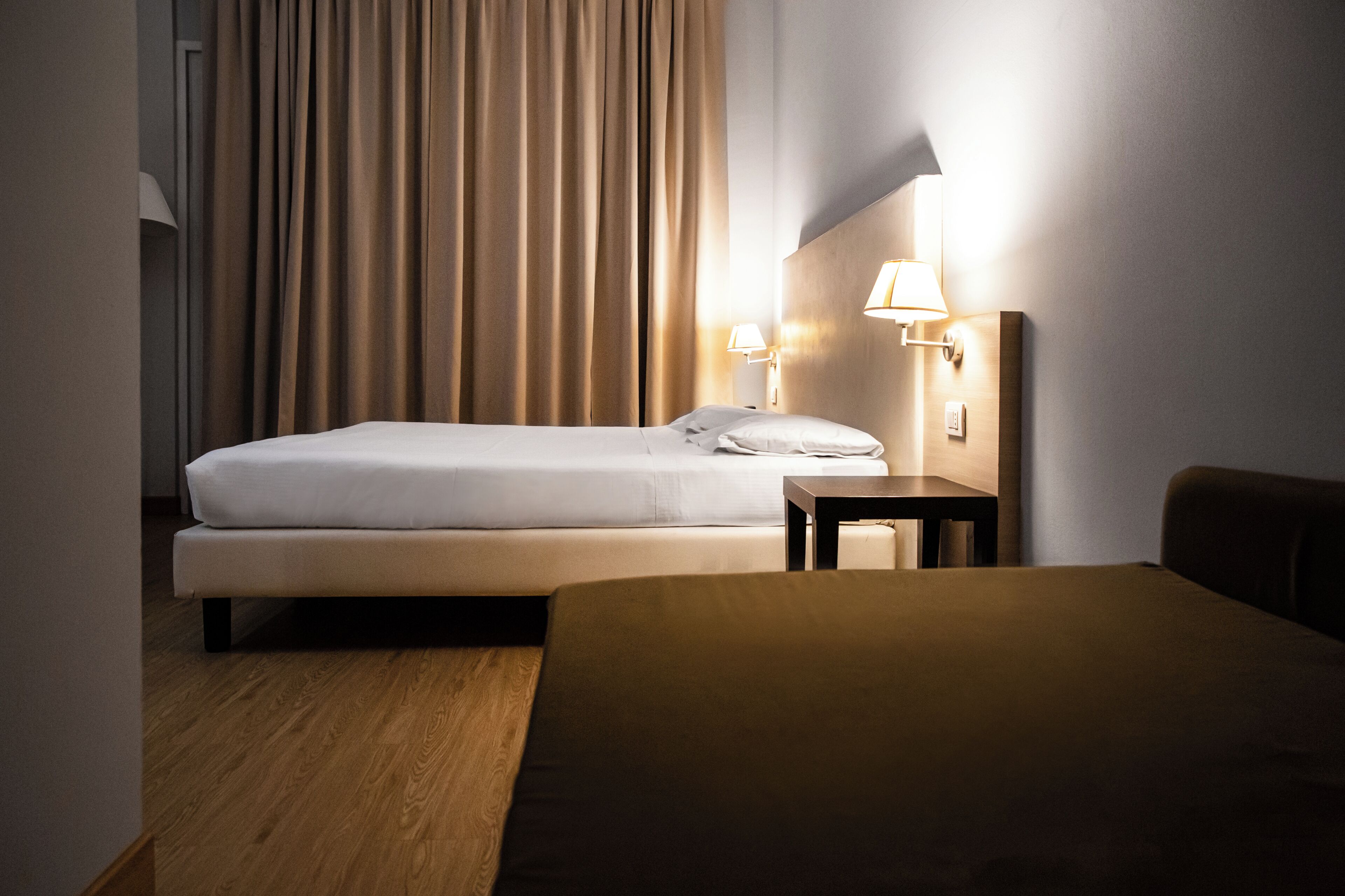 Una habitació d'hotel amb un llit, una taula de nit amb una làmpada, un capçal de llit i una cortina.