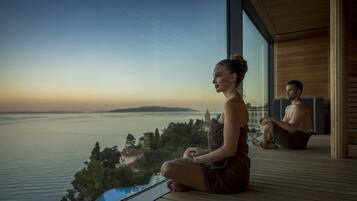 A couple meditating on a wooden deck overlooking a body of water with a distant view of a cityscape.