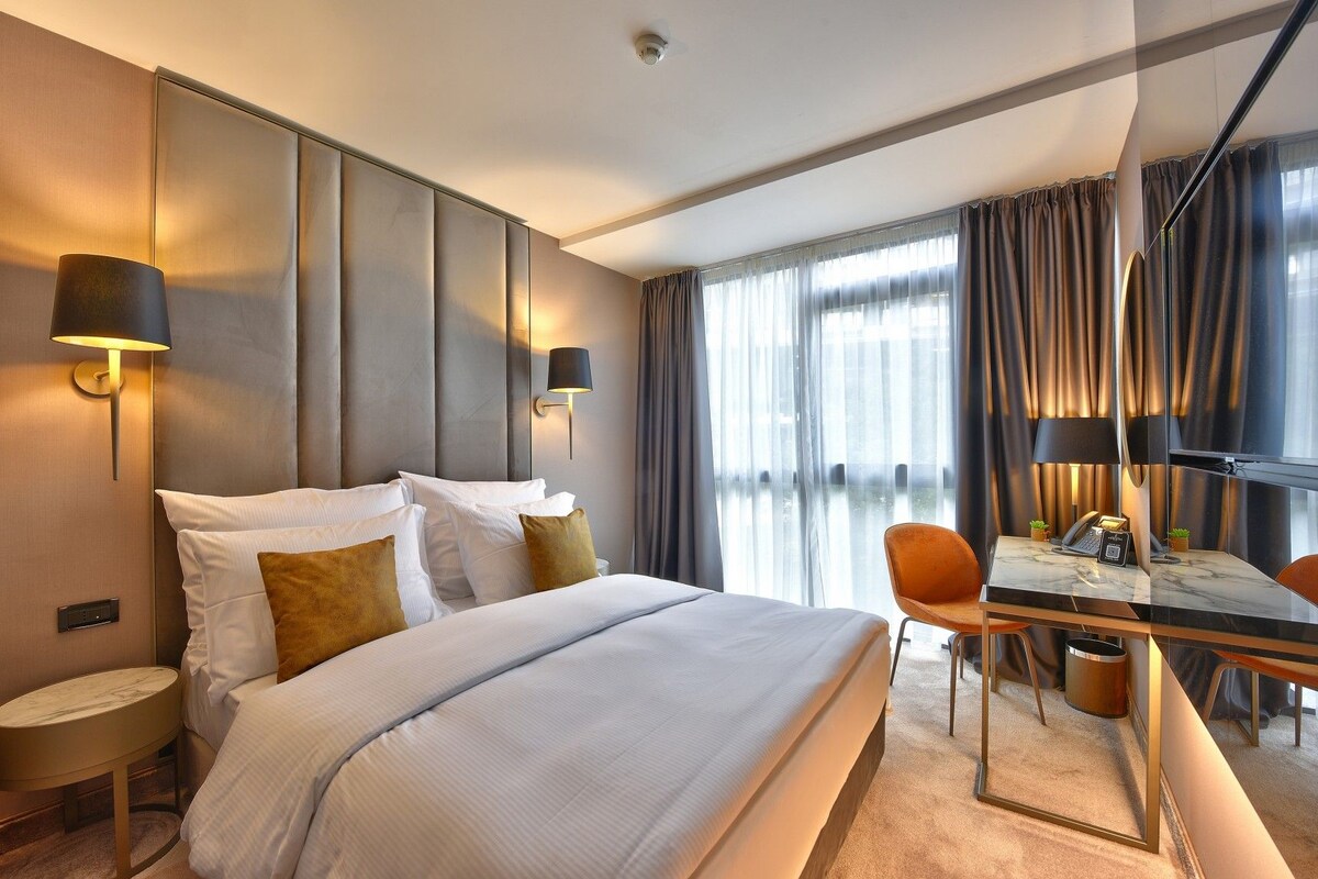 A hotel room with a large bed, two bedside lamps, a desk with a chair, and a view of the outdoors through large windows.