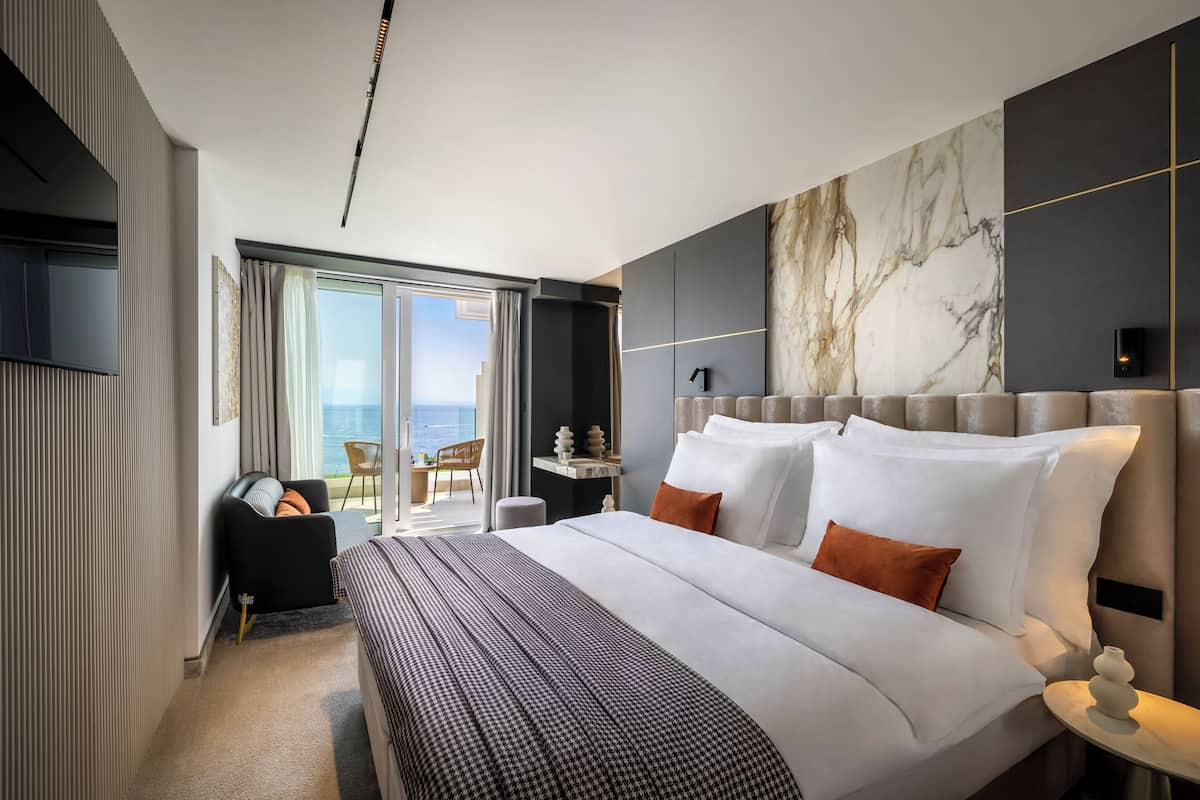 A modern hotel room with a large bed, a flat-screen TV, a marble accent wall, and a balcony with a view of the sea.