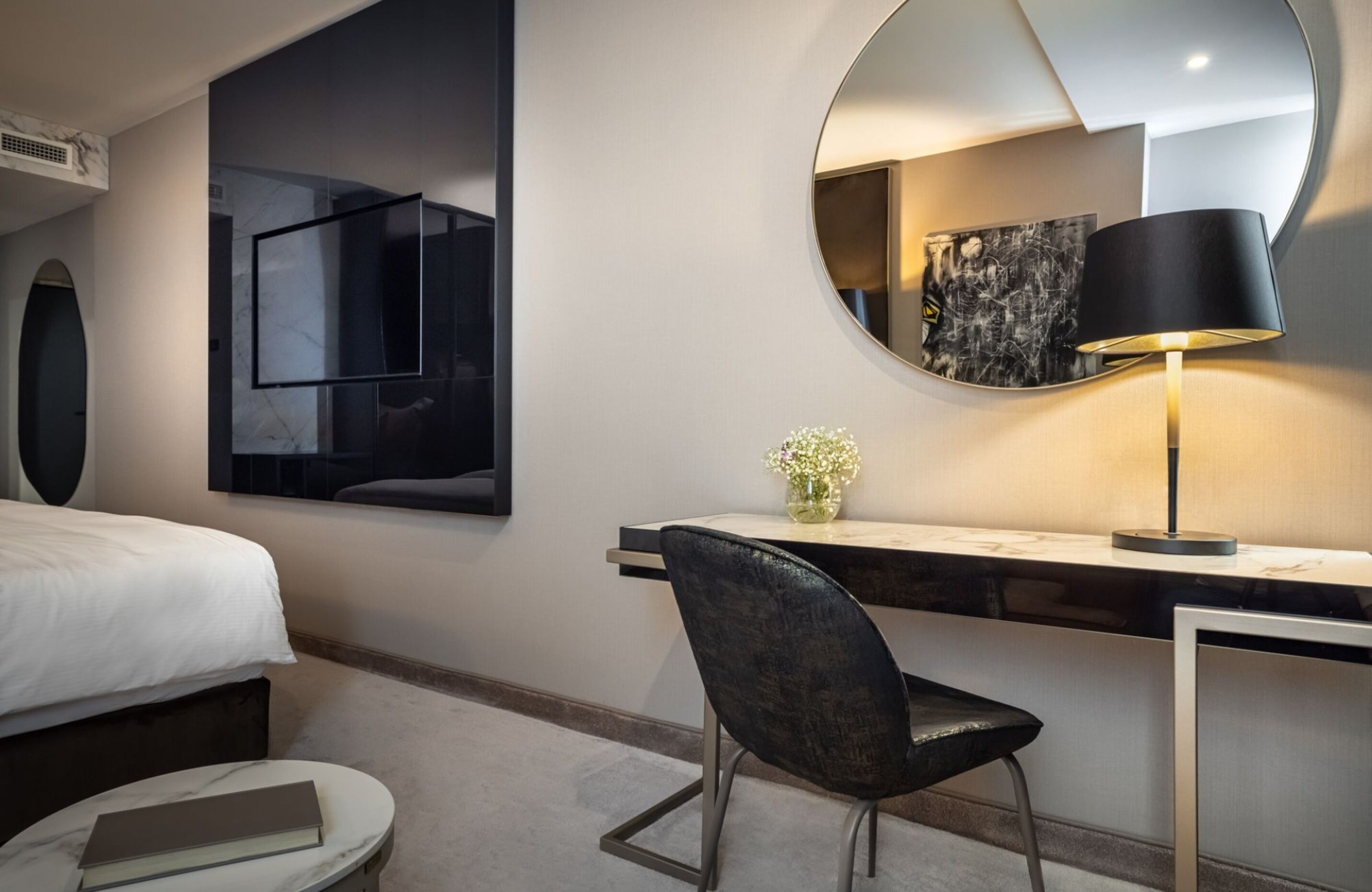 Una habitación de hotel moderna con cama, escritorio con silla, espejo redondo y lámpara.