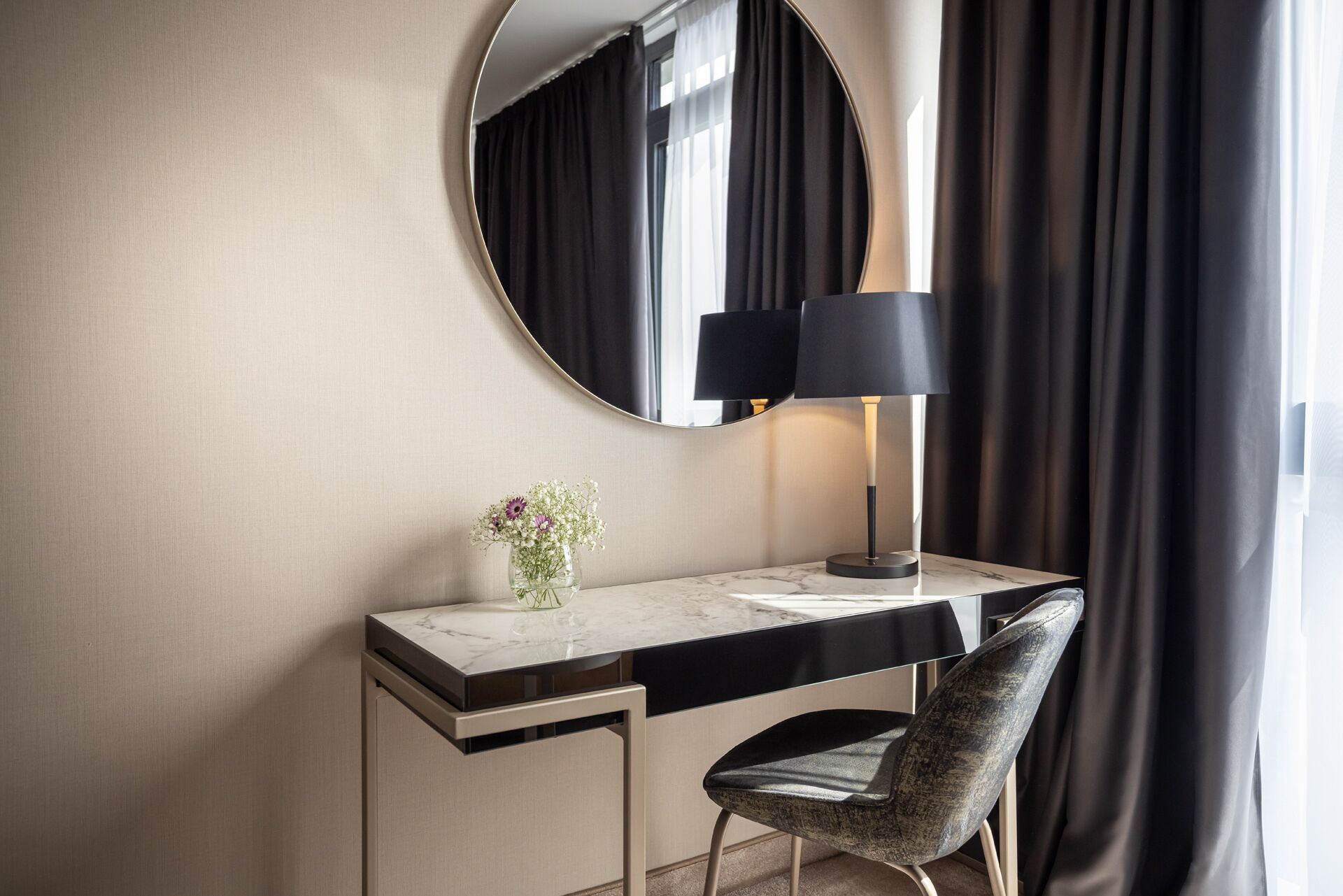 Un escritorio moderno de hotel con encimera de mármol, una lámpara negra, un jarrón con flores y una silla.
