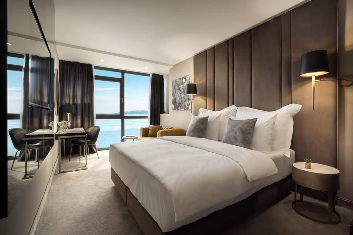 A modern hotel room with a large bed, a desk, and a view of the sea.