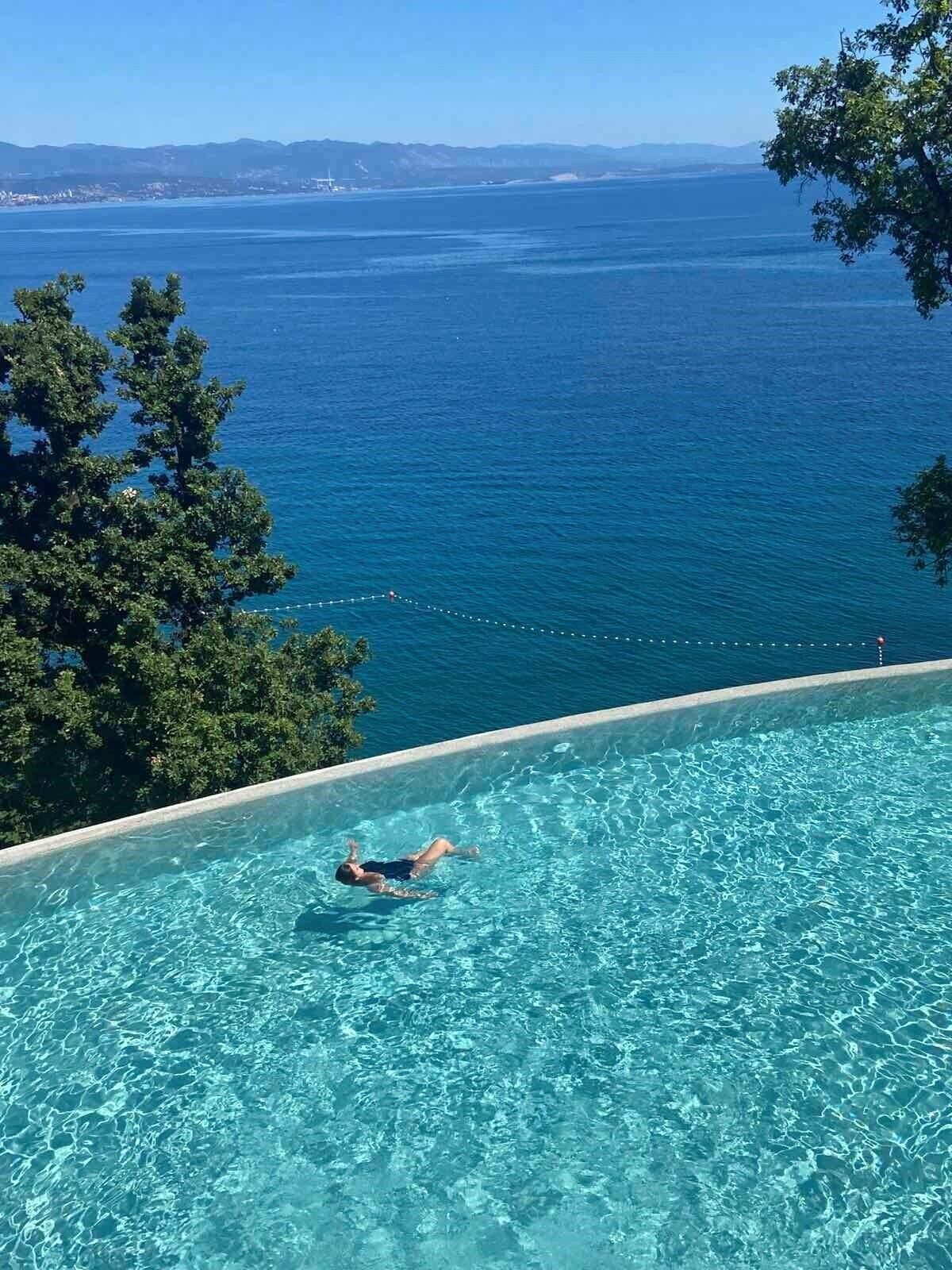 Una persona nadando en una piscina infinita azul cristalina, con vistas al océano y montañas distantes.