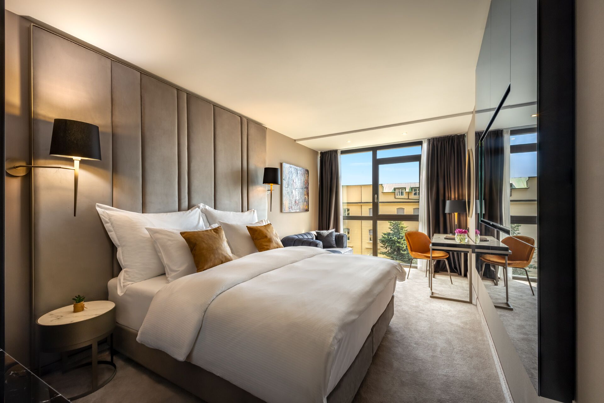 A modern hotel room with a large bed, a sitting area with a table and chairs, and a balcony with a view of the city.