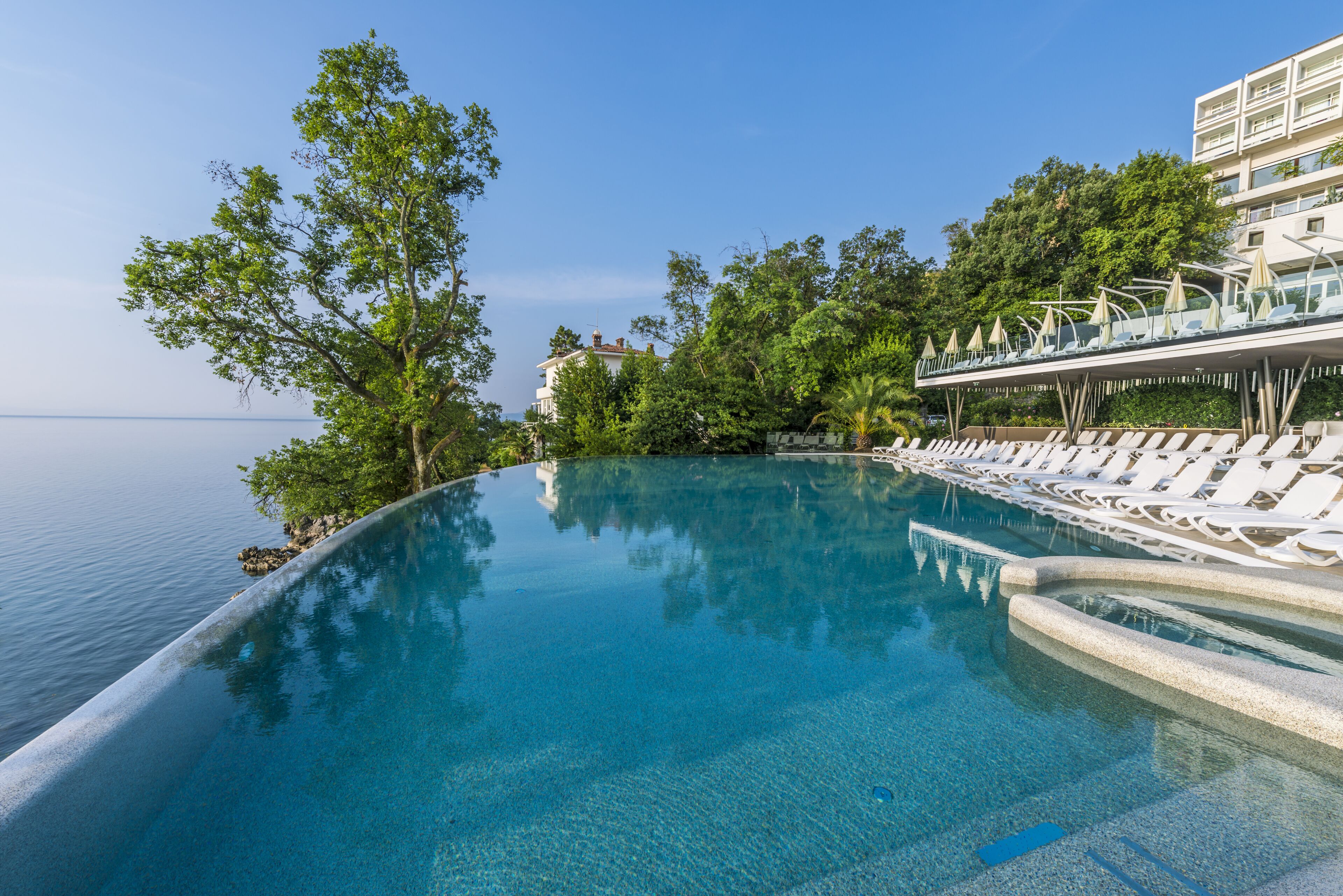 Una piscina infinita azul cristalina con sillas de descanso blancas y un puente de cristal con vistas al mar.