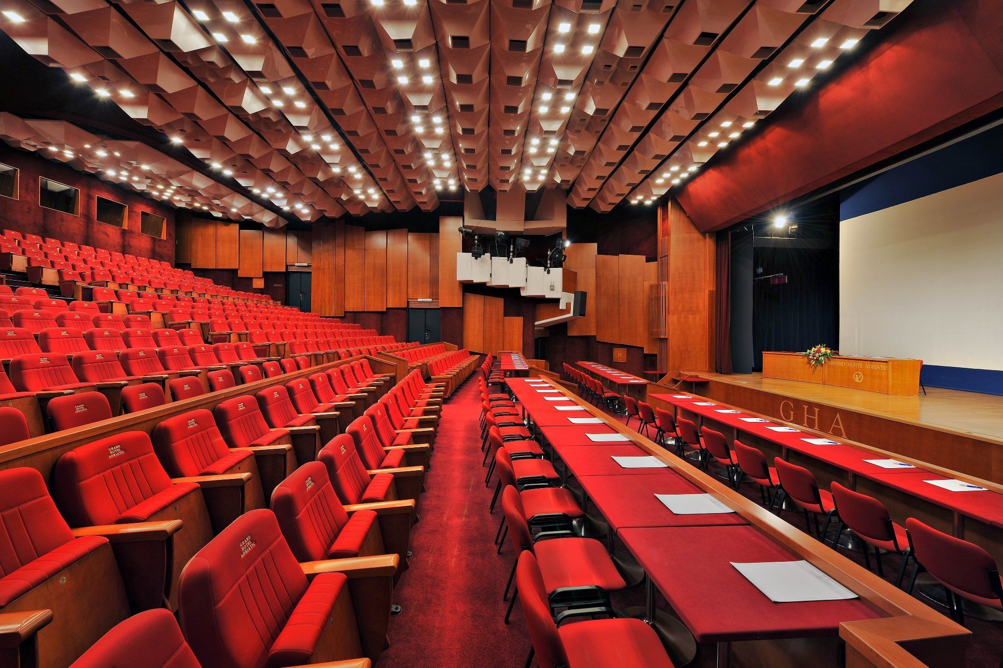 A conference room with rows of red chairs and tables, a stage with a podium, and a large screen.