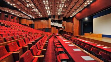 A conference room with rows of red chairs and tables, a stage with a podium, and a large screen.