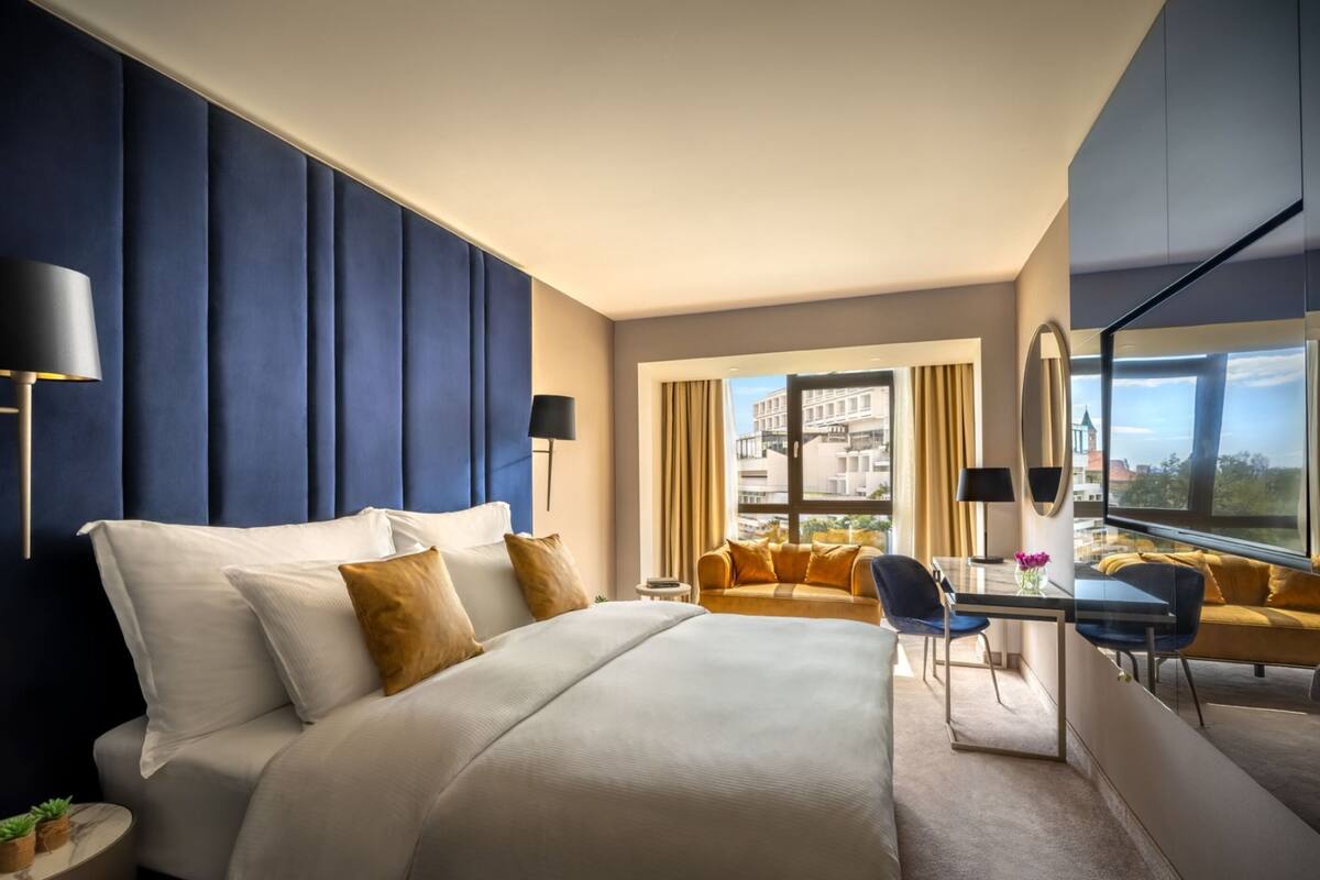 A modern hotel room with a large bed, a desk, a sofa, and a view of the city.