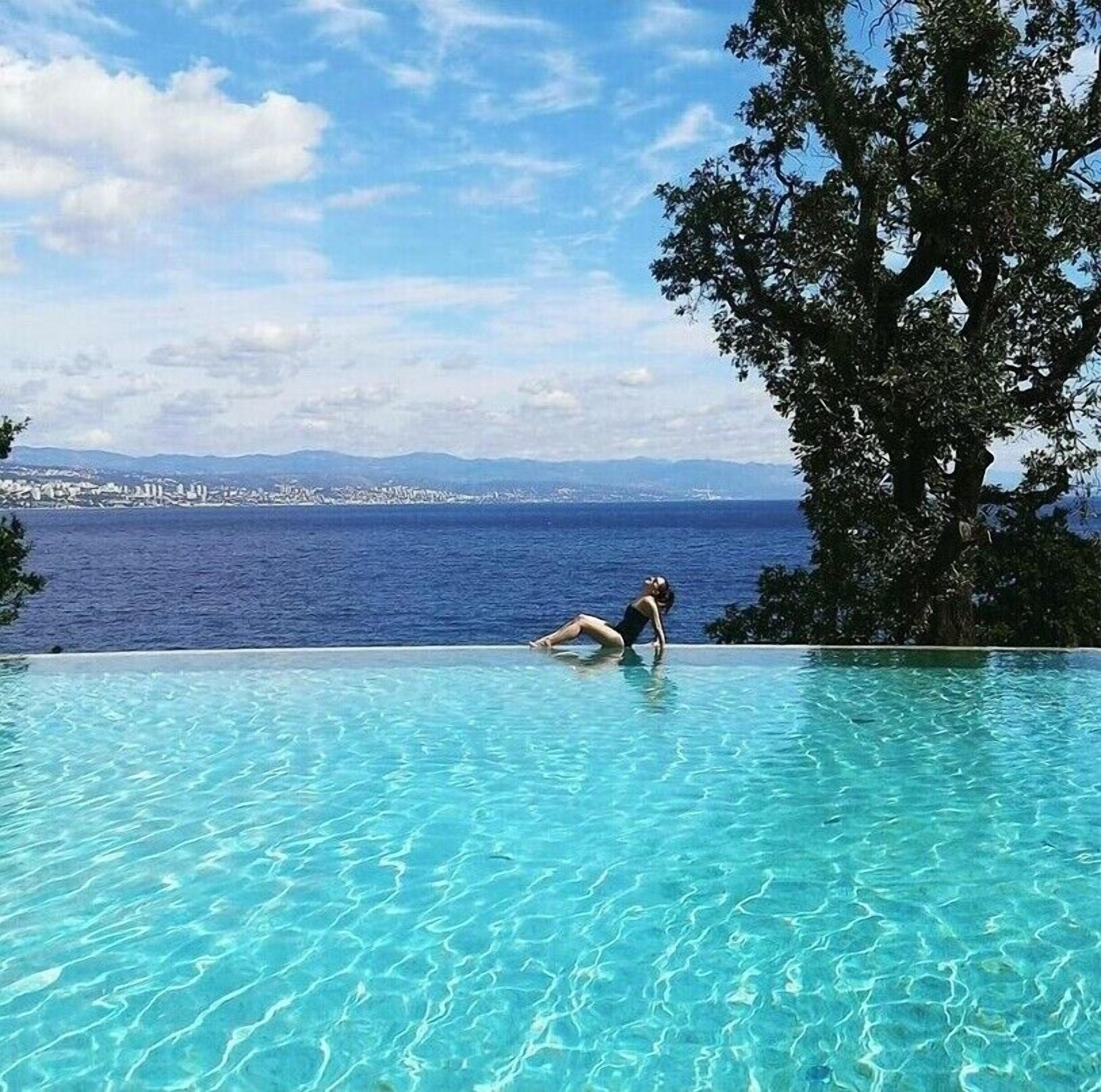A clear blue pool with a woman sitting on the edge, overlooking a coastal cityscape and the sea.