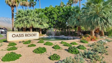 Señal de Oasis Resort rodeado por un jardín ajardinado con palmeras y un pequeño elemento acuático.