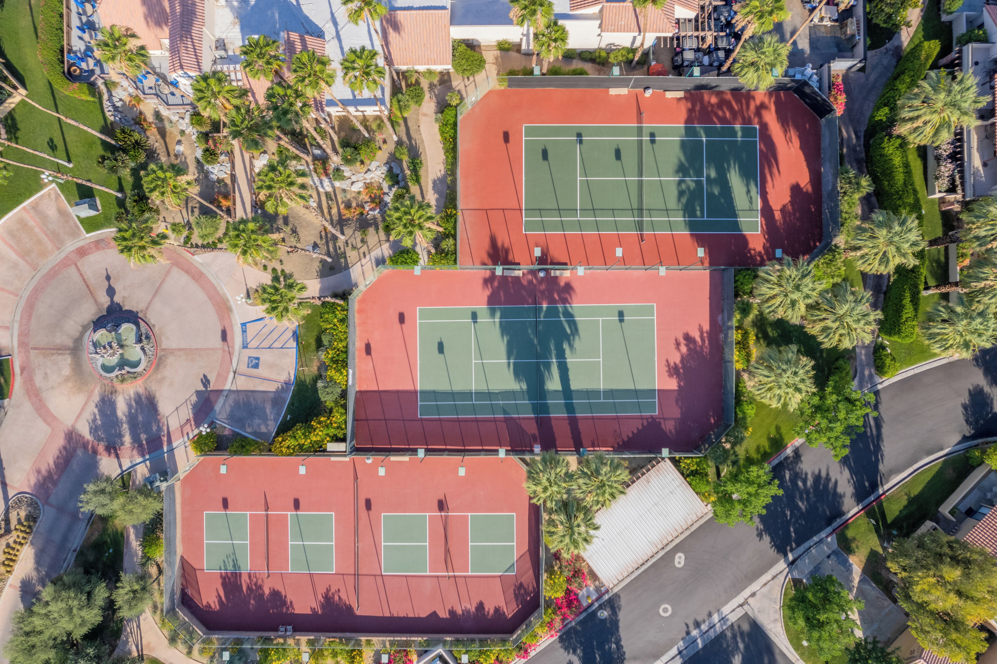 An aerial view of a sports complex with multiple tennis courts, surrounded by greenery and a parking area.