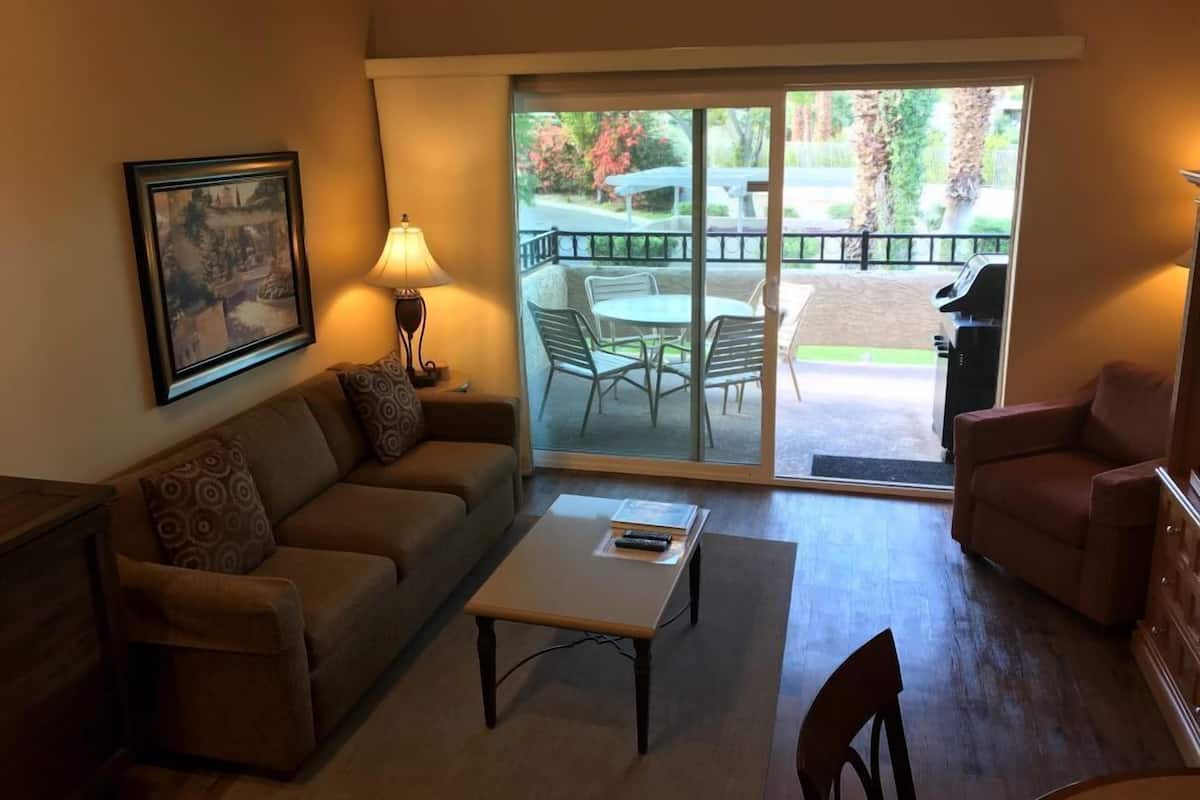 A living room with a sofa, a coffee table, and a chair. There is a sliding glass door leading to an outdoor patio with a table and chairs.