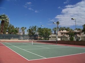An outdoor tennis court with a green surface, surrounded by a fence, palm trees, and residential buildings in the background. - The Oasis (Palm Springs)