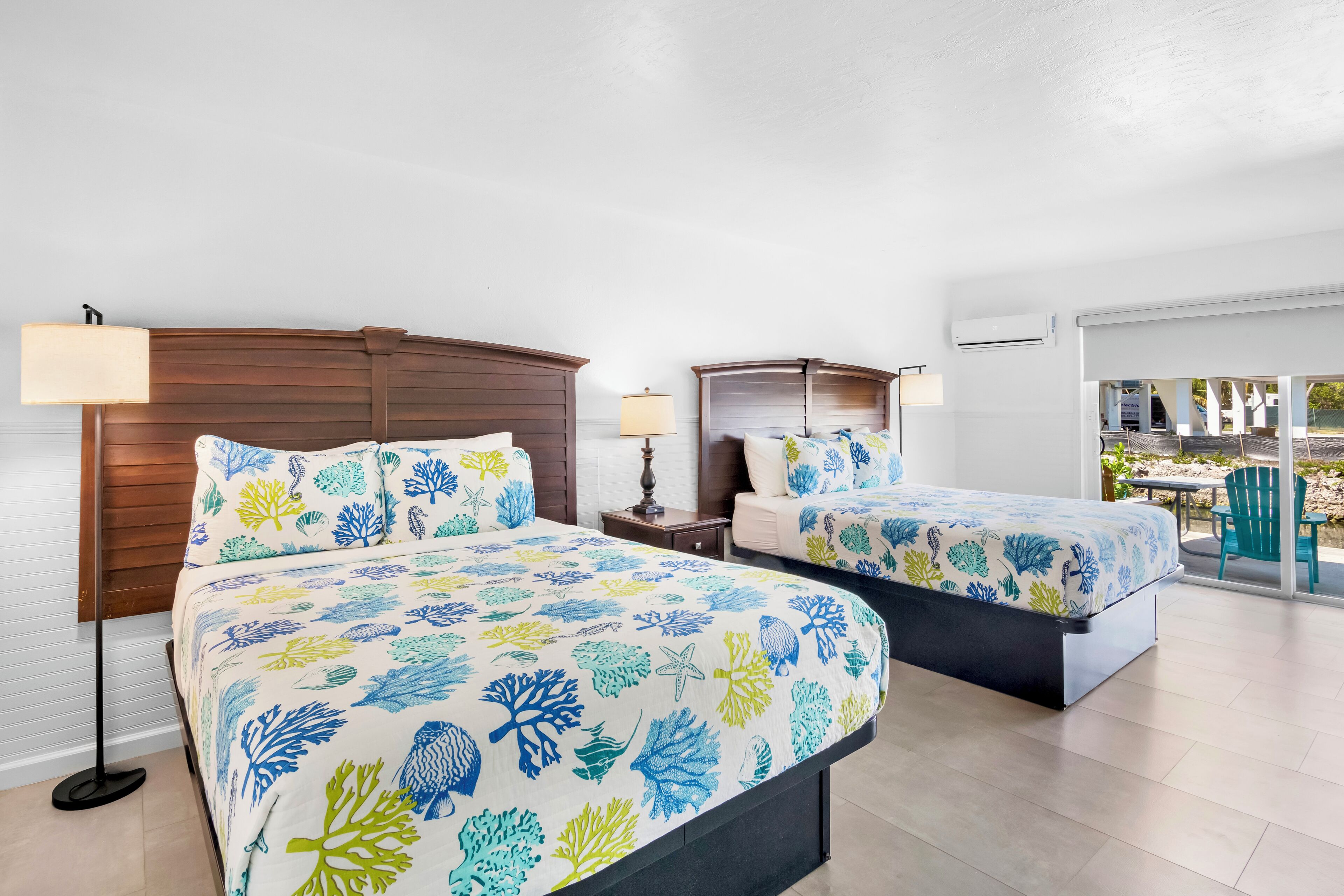 A hotel room with two beds, each with a floral bedspread, wooden headboards, bedside tables with lamps, and a view of the outdoors through a sliding glass door.