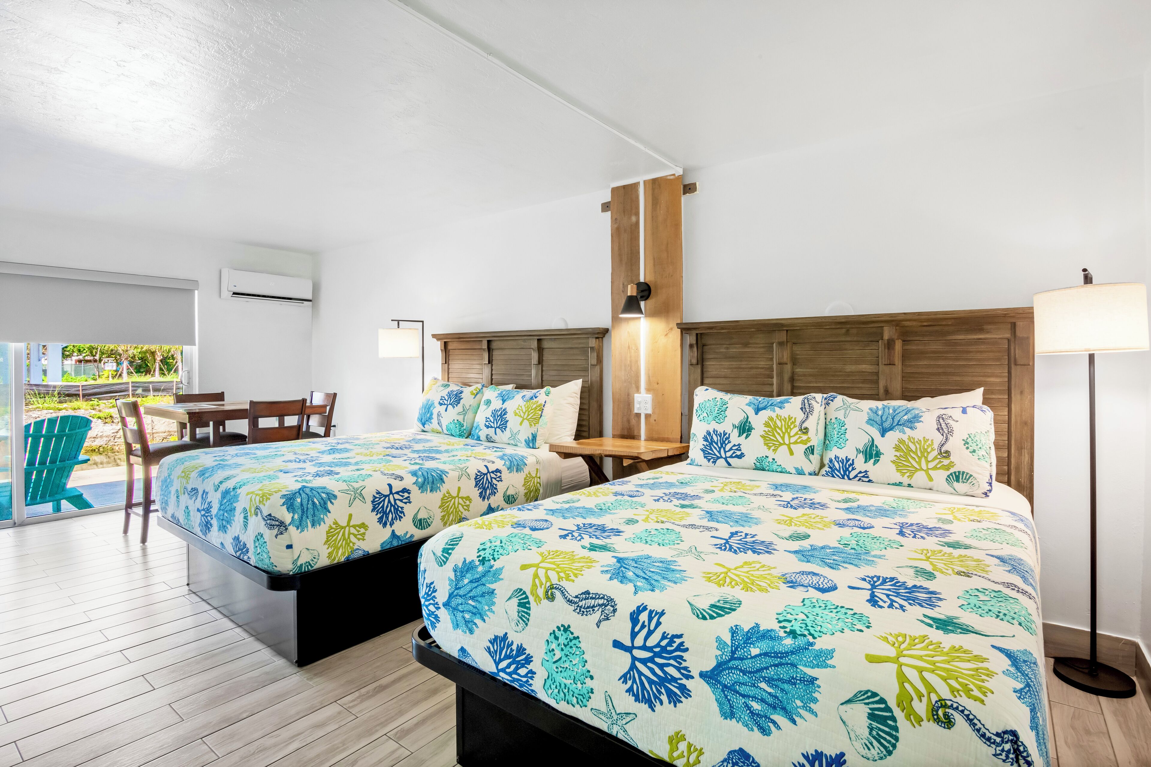 A hotel room with two beds, a wooden headboard, a table, chairs, a lamp, and a view of the outdoors.