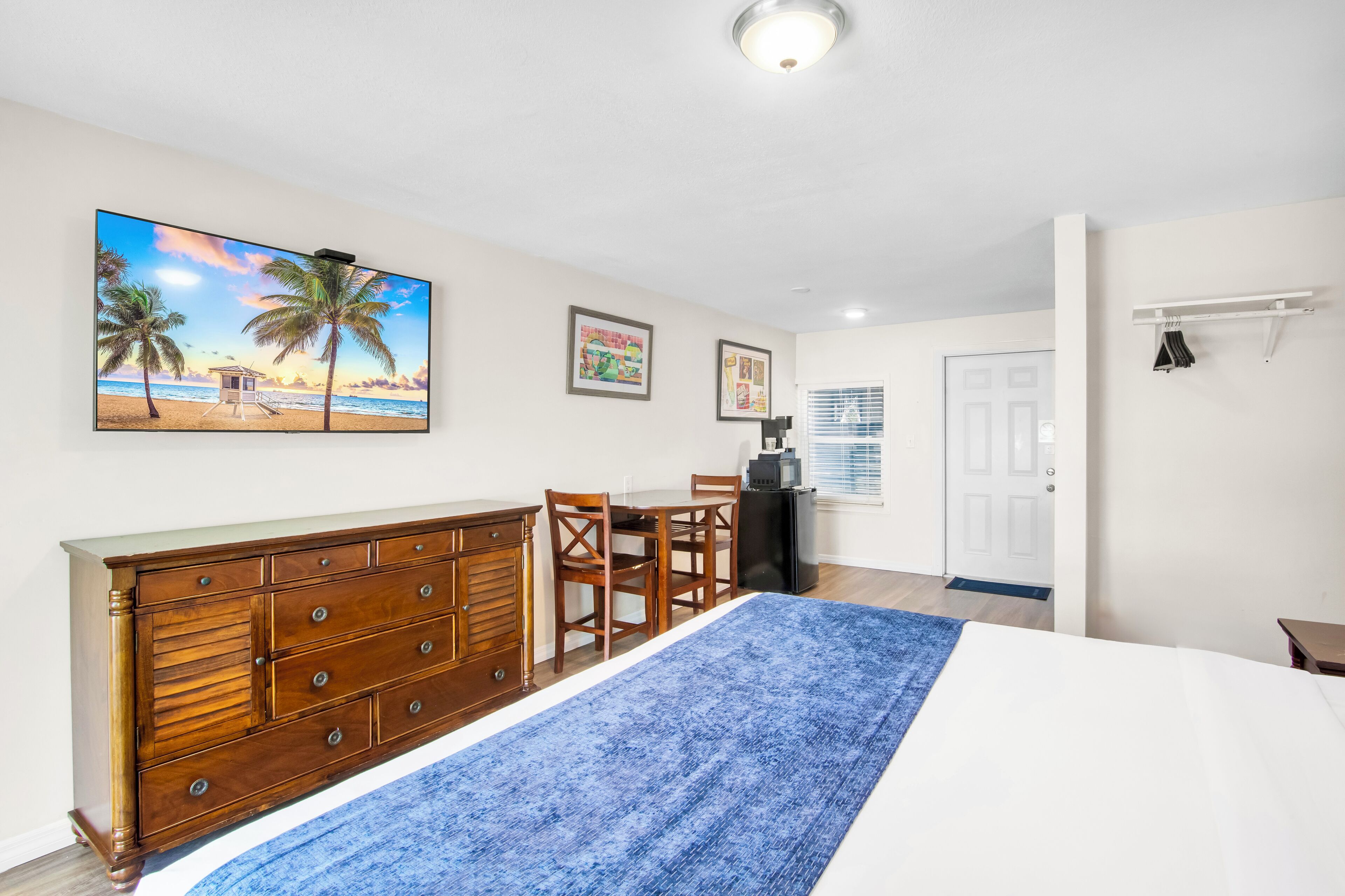 Une chambre à coucher avec un lit, une commode, un bureau et des chaises. Un tableau représentant une scène de plage est accroché au mur.