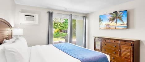 A hotel room with a bed, a dresser, a painting of a beach scene, and a view of a garden through the window.
